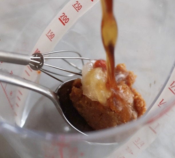 計量カップに入った合わせみそ、豆板醤、すりおろしにんにくに、めんつゆ（ストレート）が注がれている様子。豚バラもやしの味噌だれそうめんのタレ作りの初期工程。