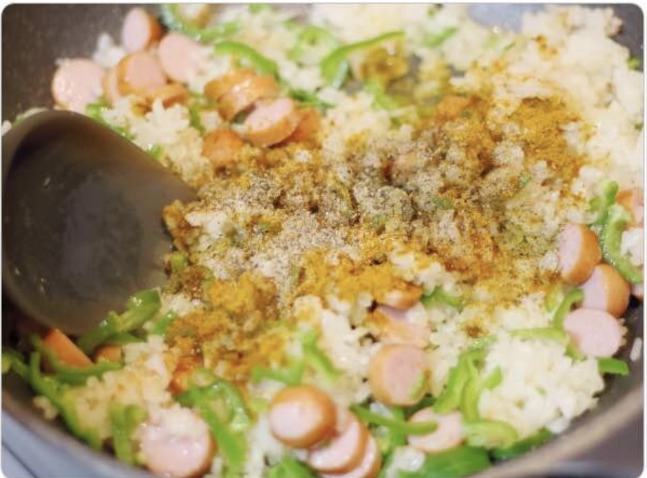 黒いフライパンの中のチャーハンに、カレー粉と塩こしょうなどのスパイスが加えられている。マヨネーズカレーチャーハンの味付け工程。