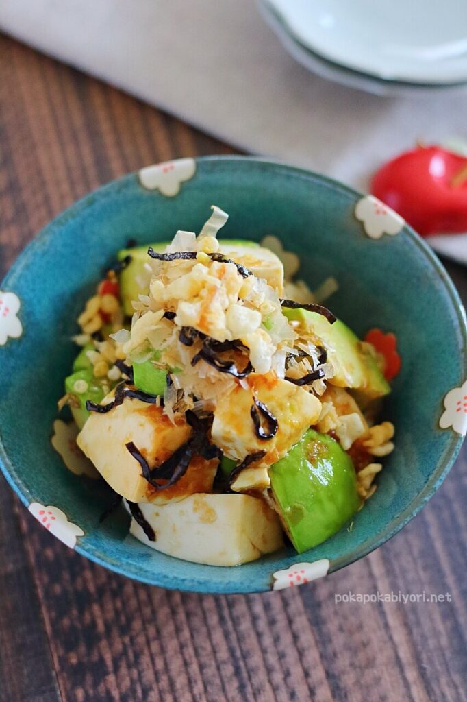 盛り付けられたアボカドと豆腐の和え物。削り節、塩昆布、天かすがトッピングされ、青い小鉢に入っている。背景に赤い小物。