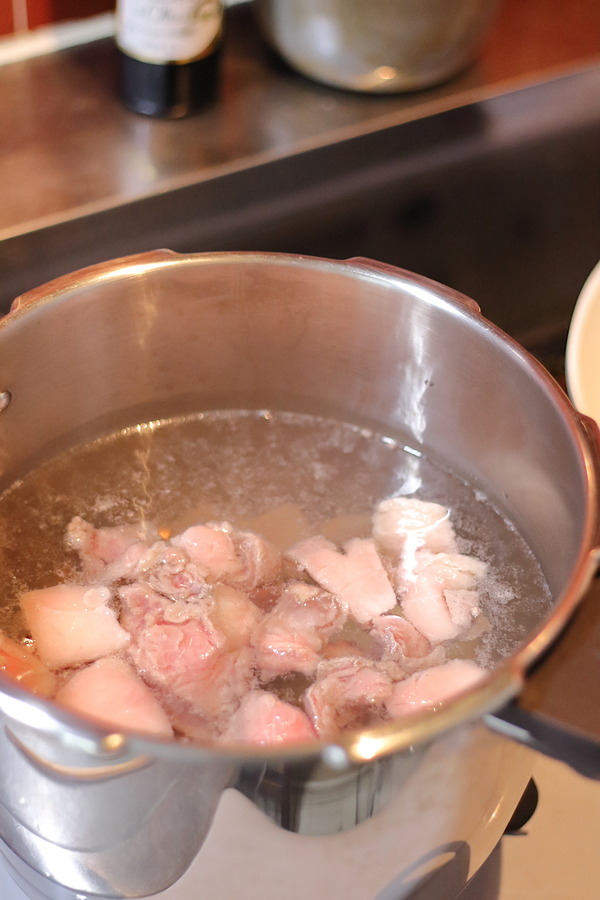 圧力鍋の中で、食べやすい大きさに切った牛すじ肉をたっぷりの水で下茹でしている様子。沸騰してアクが浮いており、臭み取りの重要な工程