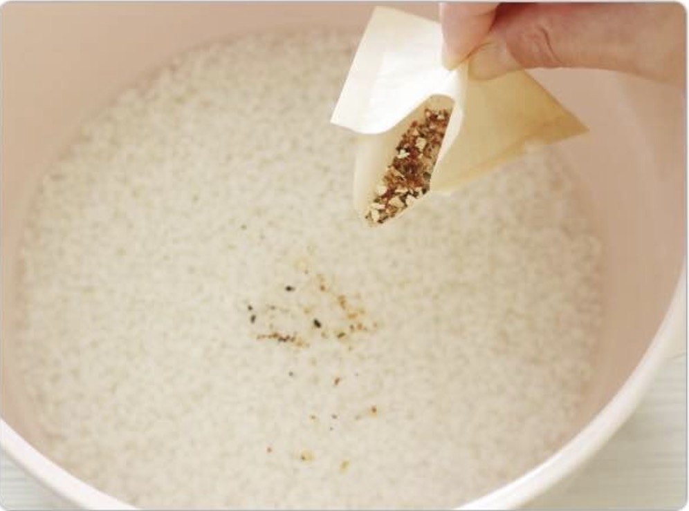 ピンクの鍋に入った洗った白米に、だしパックから粉末だしを振り入れている様子。