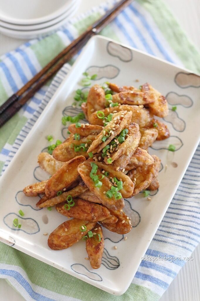 甘辛く揚げ焼きにしたごぼうに白ごまと小口切りの万能ねぎを散らした和食の副菜。白い長皿に盛り付けられ、箸が添えられている。