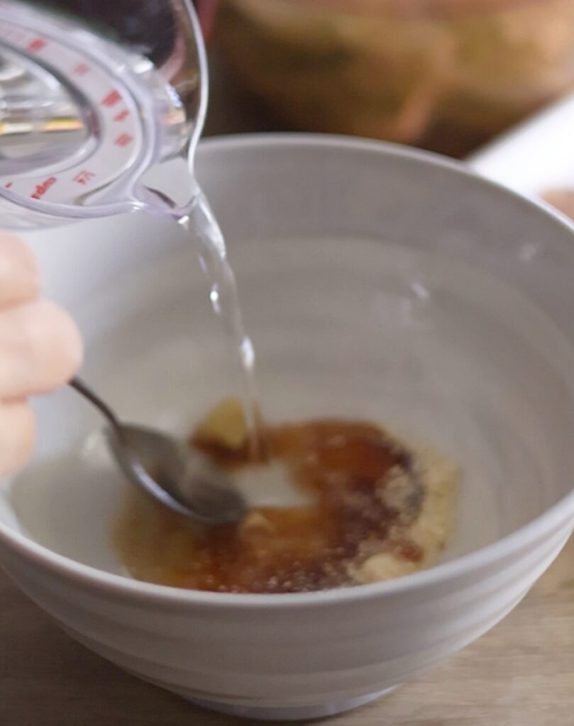 豚バラ旨塩うどんの作り方。うどん鉢に入れた鶏ガラや醤油などの調味料に水を加えてスープベースを作る工程