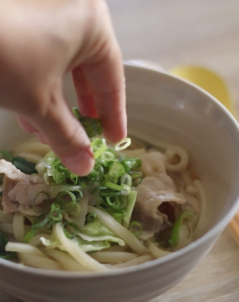 完成した豚バラ旨塩うどんの仕上げ。豚肉やキャベツと混ぜ合わせたうどんの上に、刻みねぎをトッピングしている盛り付けシーン