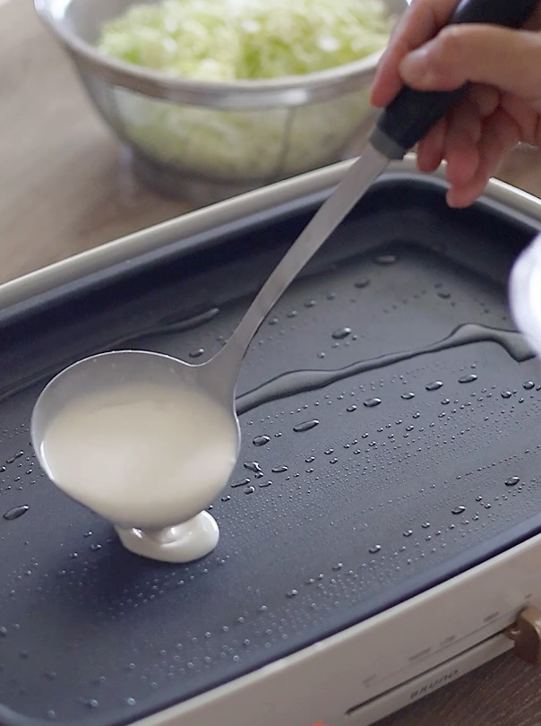 180°Cに熱したホットプレートにサラダ油を引き、生地を円形に薄く広げ、上に削り節を振り、キャベツ、ねぎ、揚げ玉、紅生姜、豚肉の順に重ねます。